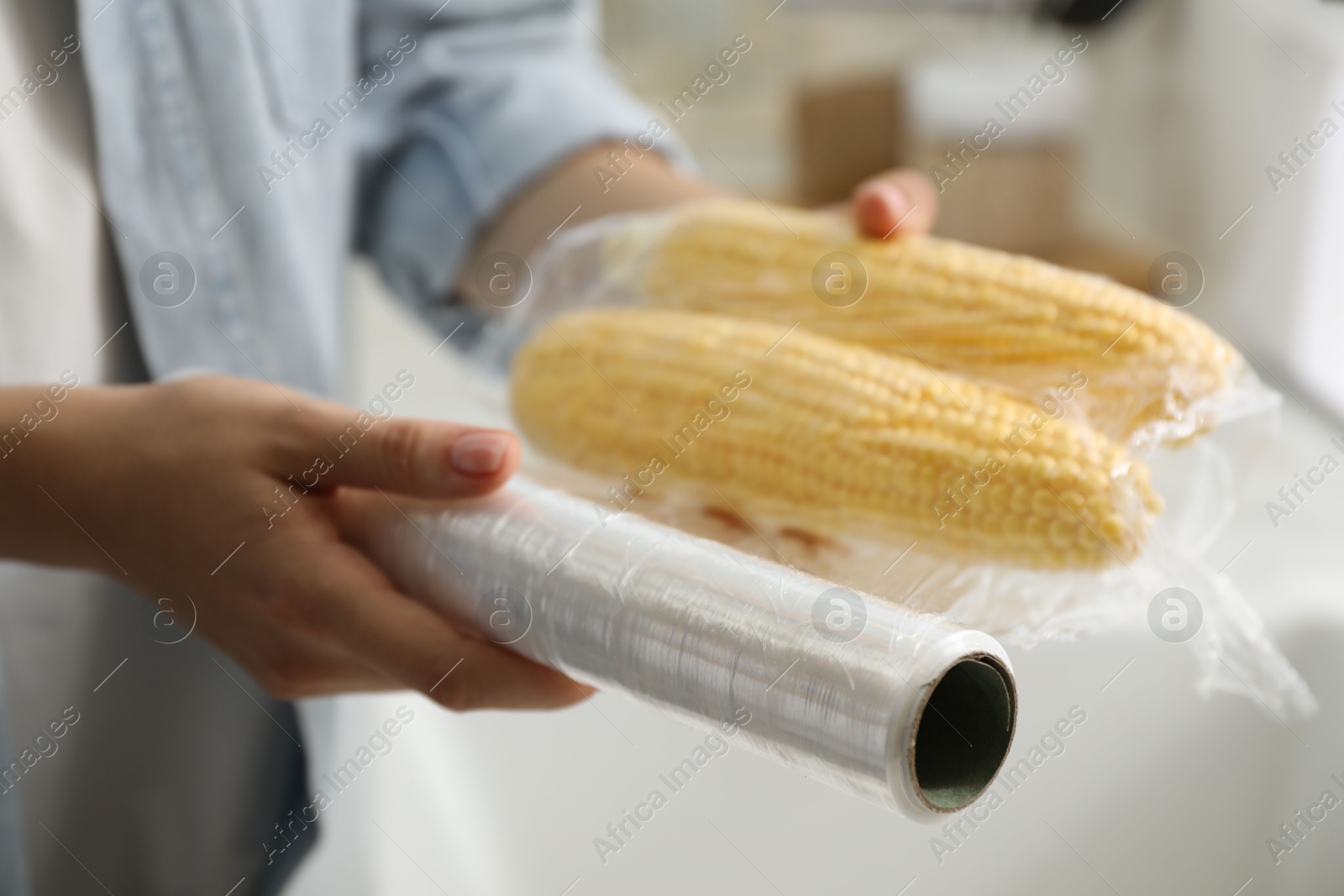 Photo of Woman putting plastic food wrap over corncobs in kitchen, closeup