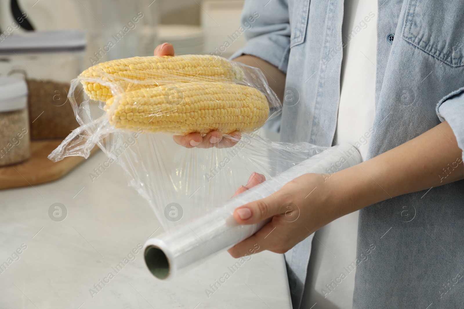 Photo of Woman putting plastic food wrap over corncobs at countertop in kitchen, closeup