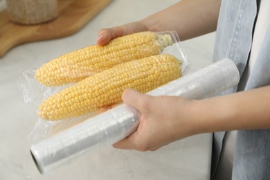 Woman putting plastic food wrap over corncobs in kitchen, closeup