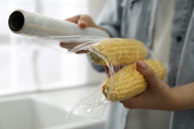 Woman putting plastic food wrap over corncobs in kitchen, closeup