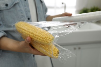 Woman putting plastic food wrap over corncobs in kitchen, closeup