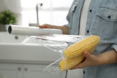 Woman putting plastic food wrap over corncobs in kitchen, closeup