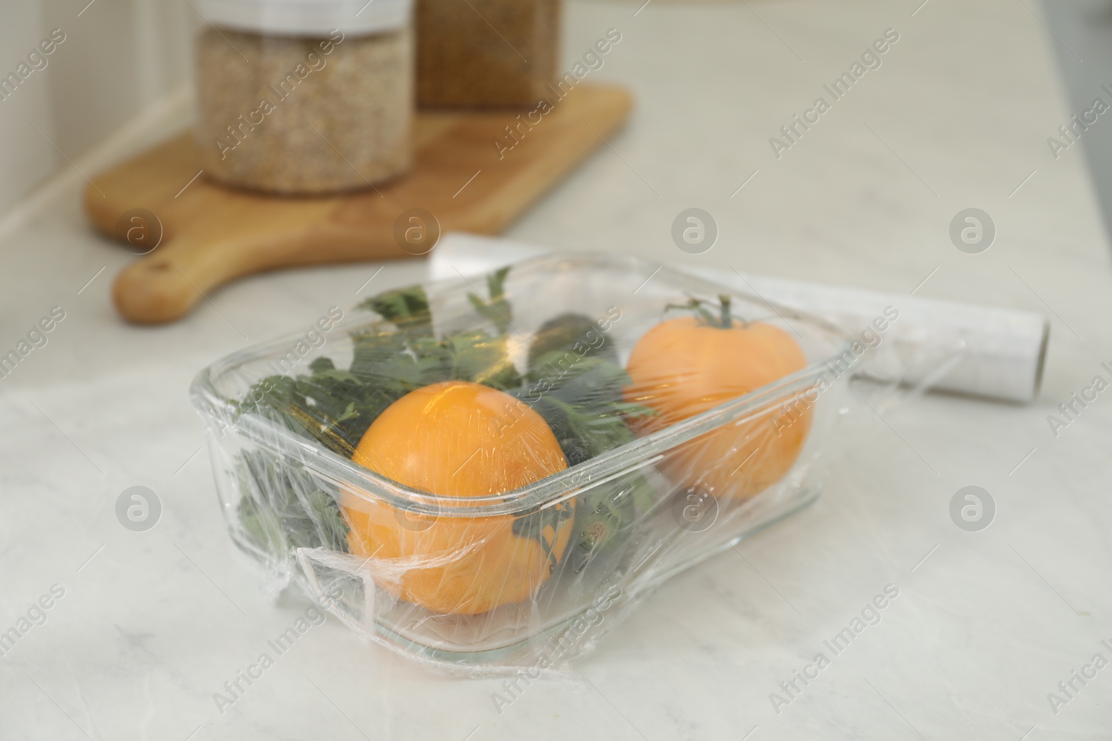 Photo of Glass container of fresh vegetables with plastic food wrap on white marble table, closeup