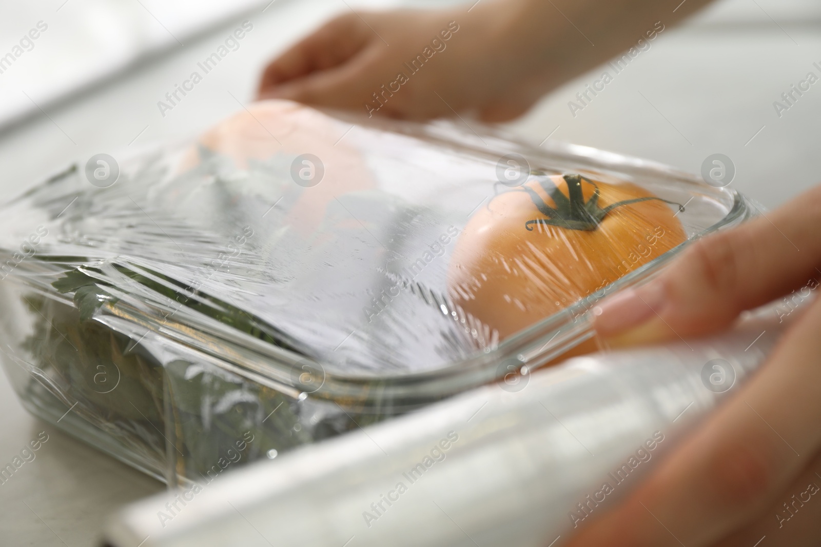 Photo of Woman putting plastic food wrap over glass container with vegetables at countertop in kitchen, closeup