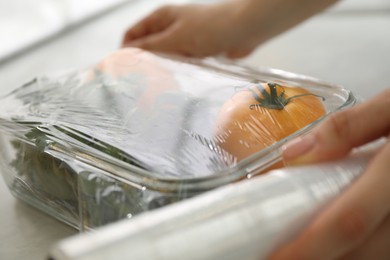 Woman putting plastic food wrap over glass container with vegetables at countertop in kitchen, closeup