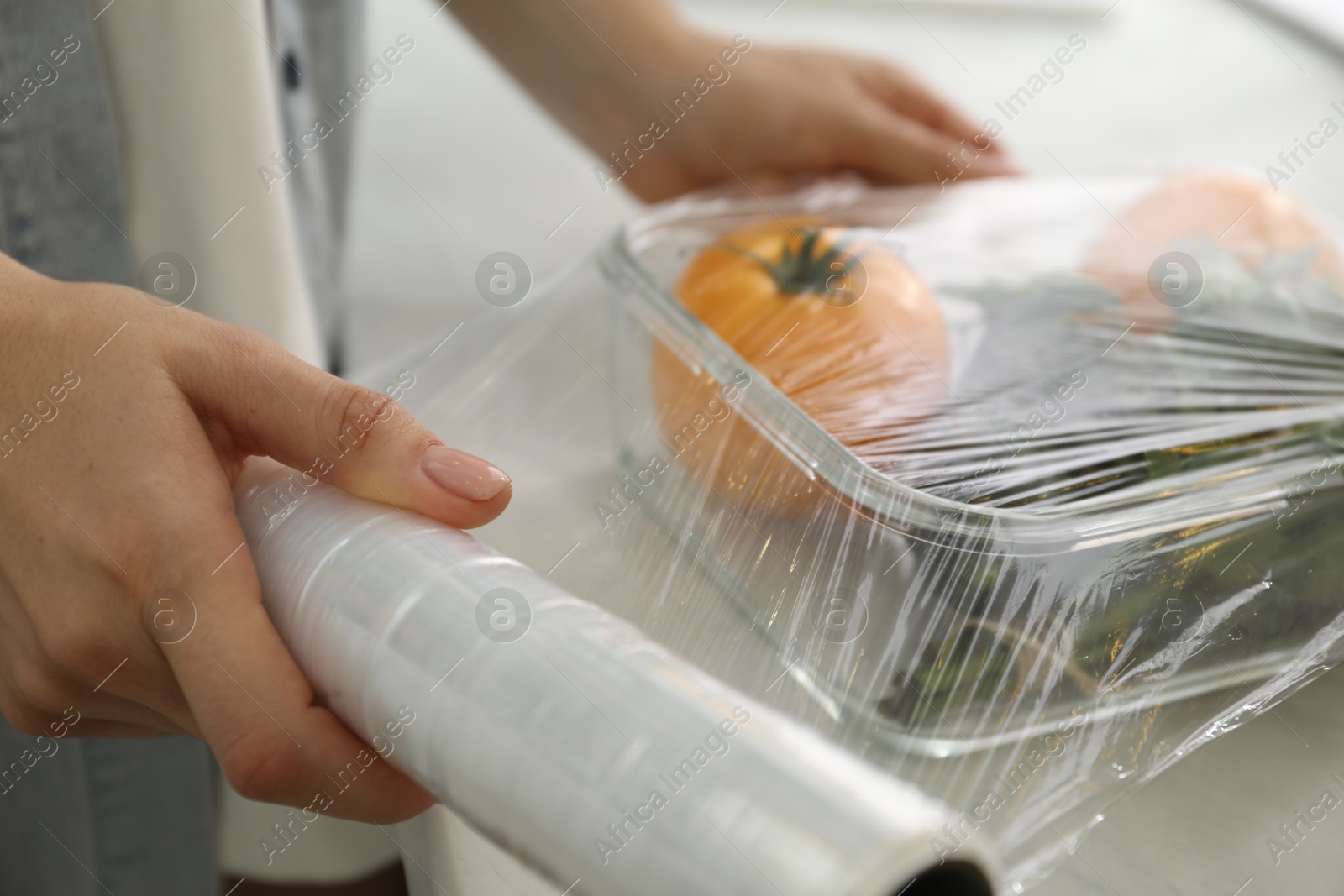 Photo of Woman putting plastic food wrap over glass container with vegetables at countertop in kitchen, closeup