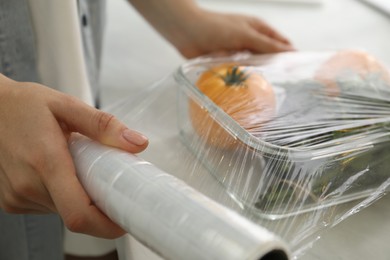 Woman putting plastic food wrap over glass container with vegetables at countertop in kitchen, closeup