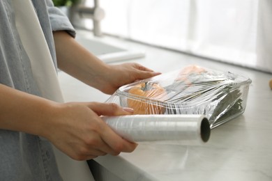 Photo of Woman putting plastic food wrap over glass container with vegetables at countertop in kitchen, closeup