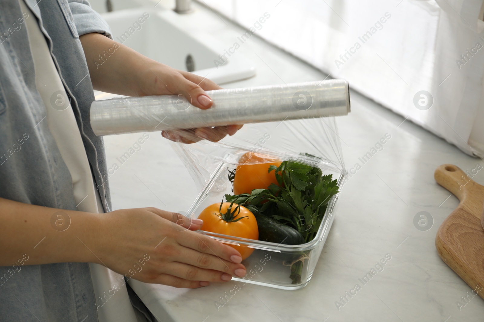 Photo of Woman putting plastic food wrap over glass container with vegetables at countertop in kitchen, closeup