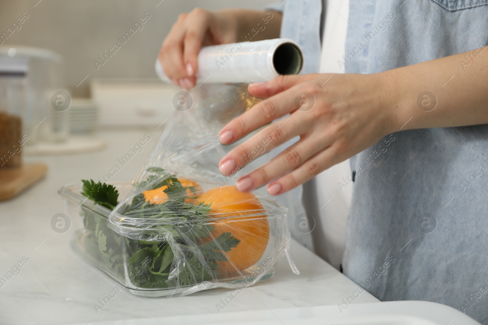 Photo of Woman putting plastic food wrap over glass container with vegetables at countertop in kitchen, closeup