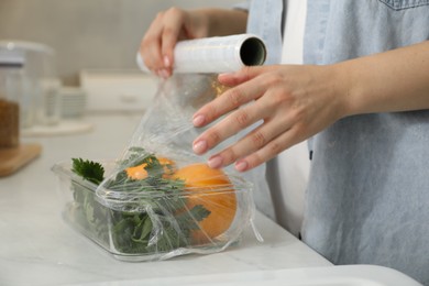 Woman putting plastic food wrap over glass container with vegetables at countertop in kitchen, closeup