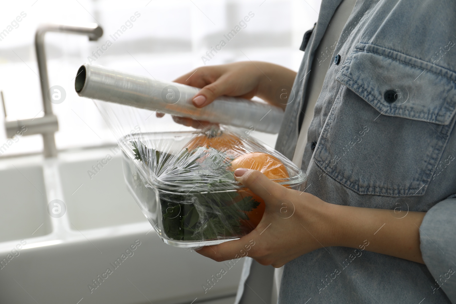 Photo of Woman putting plastic food wrap over glass container with vegetables in kitchen, closeup