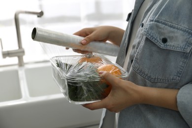 Woman putting plastic food wrap over glass container with vegetables in kitchen, closeup