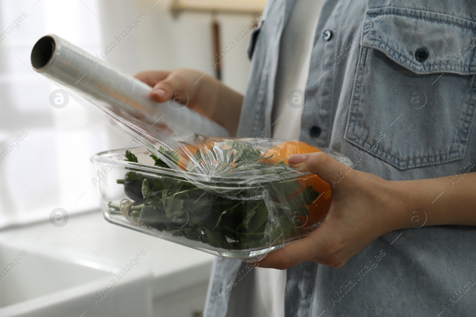 Photo of Woman putting plastic food wrap over glass container with vegetables in kitchen, closeup