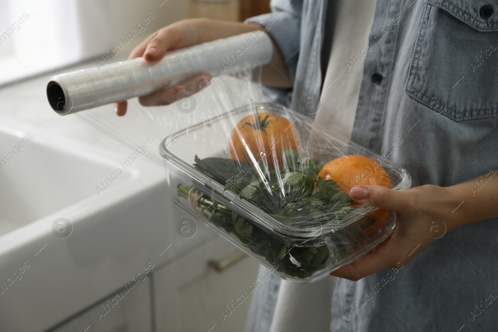 Photo of Woman putting plastic food wrap over glass container with vegetables in kitchen, closeup