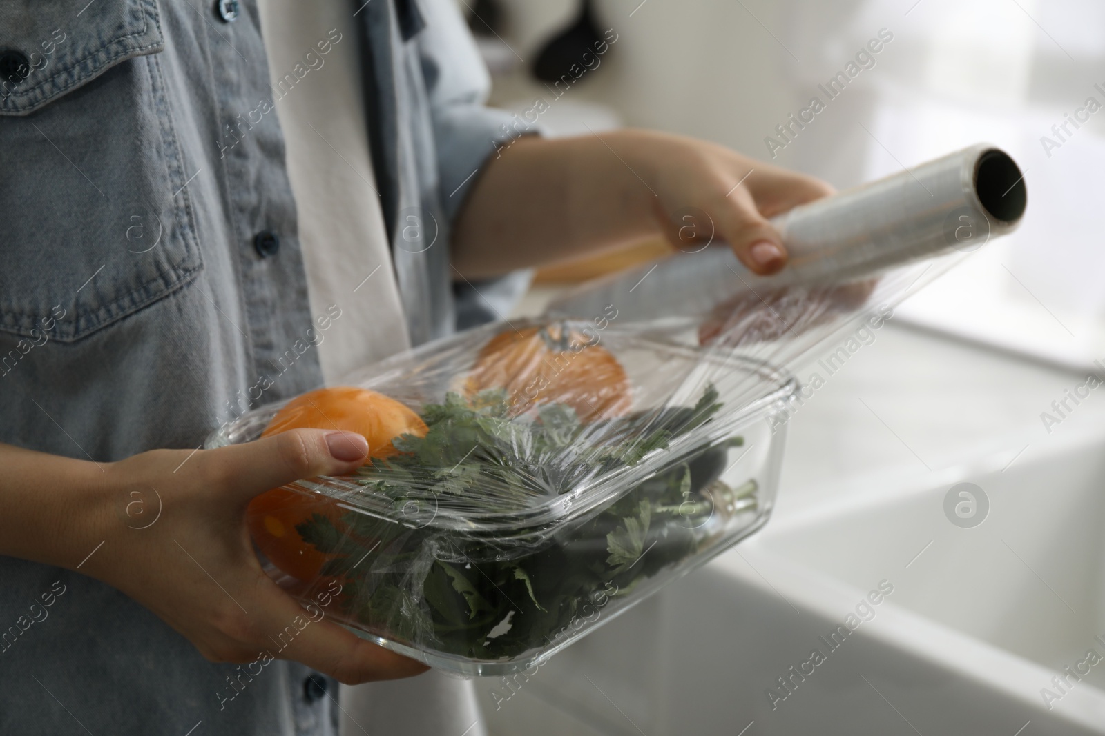 Photo of Woman putting plastic food wrap over glass container with vegetables in kitchen, closeup