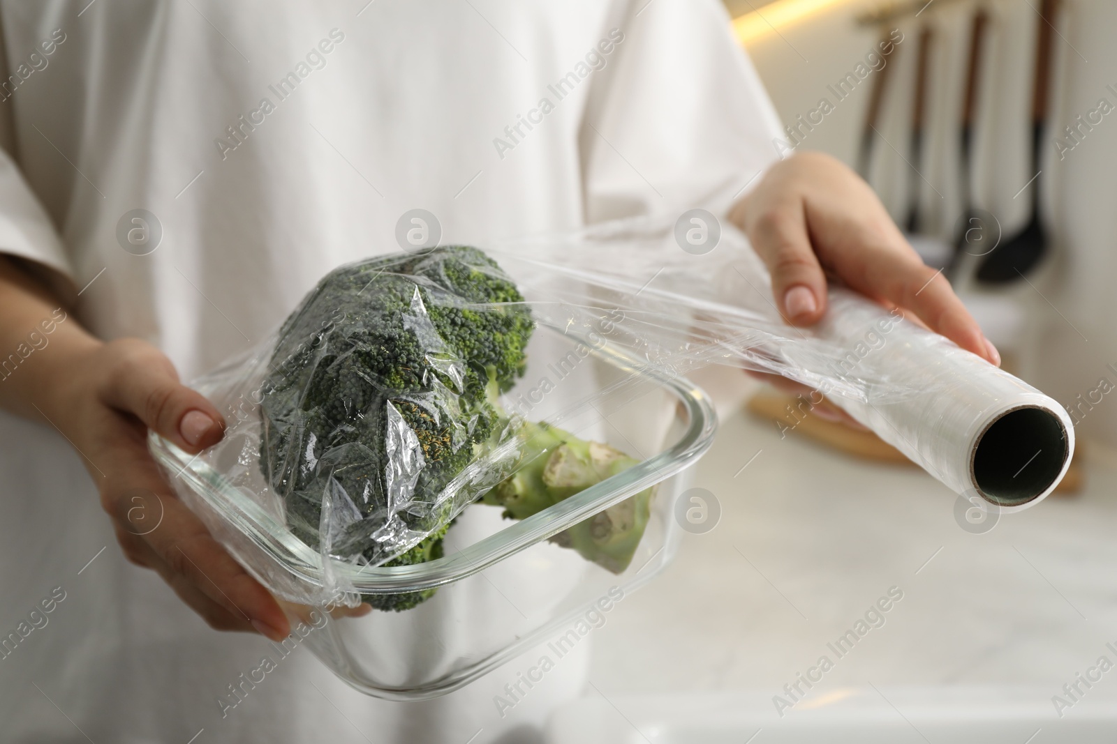 Photo of Woman putting plastic food wrap over glass container with broccoli in kitchen, closeup