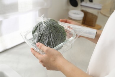 Photo of Woman putting plastic food wrap over glass container with broccoli in kitchen, closeup