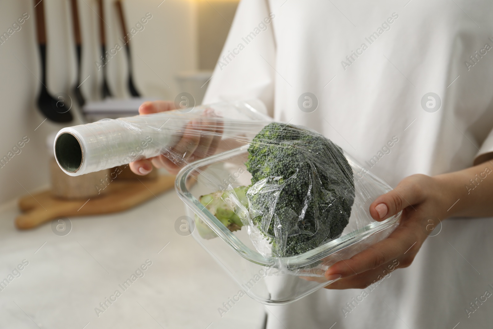 Photo of Woman putting plastic food wrap over glass container with broccoli in kitchen, closeup