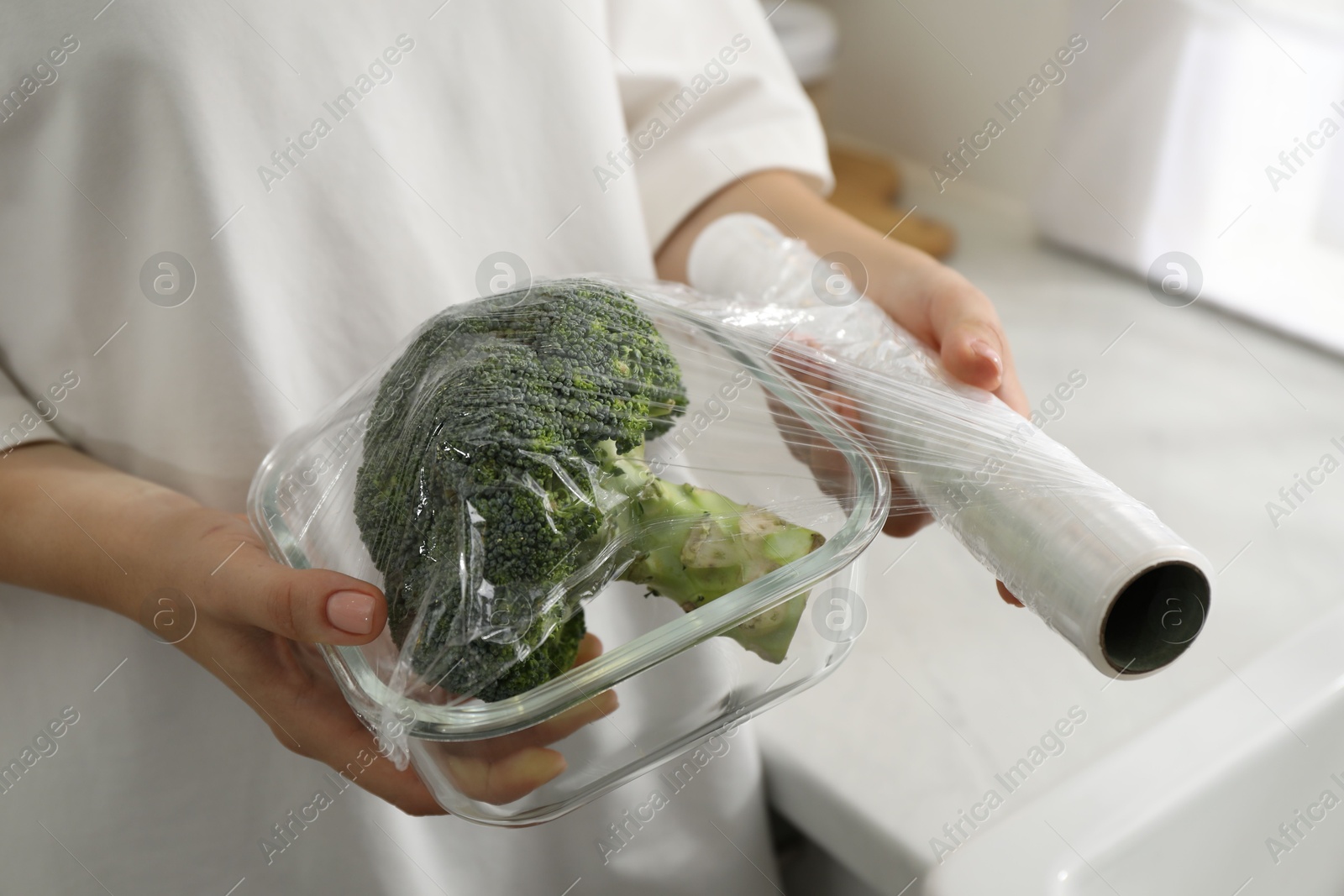 Photo of Woman putting plastic food wrap over glass container with broccoli in kitchen, closeup