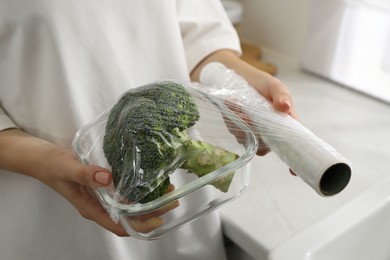 Woman putting plastic food wrap over glass container with broccoli in kitchen, closeup