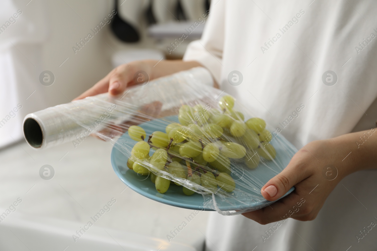 Photo of Woman putting plastic food wrap over plate with grapes in kitchen, closeup