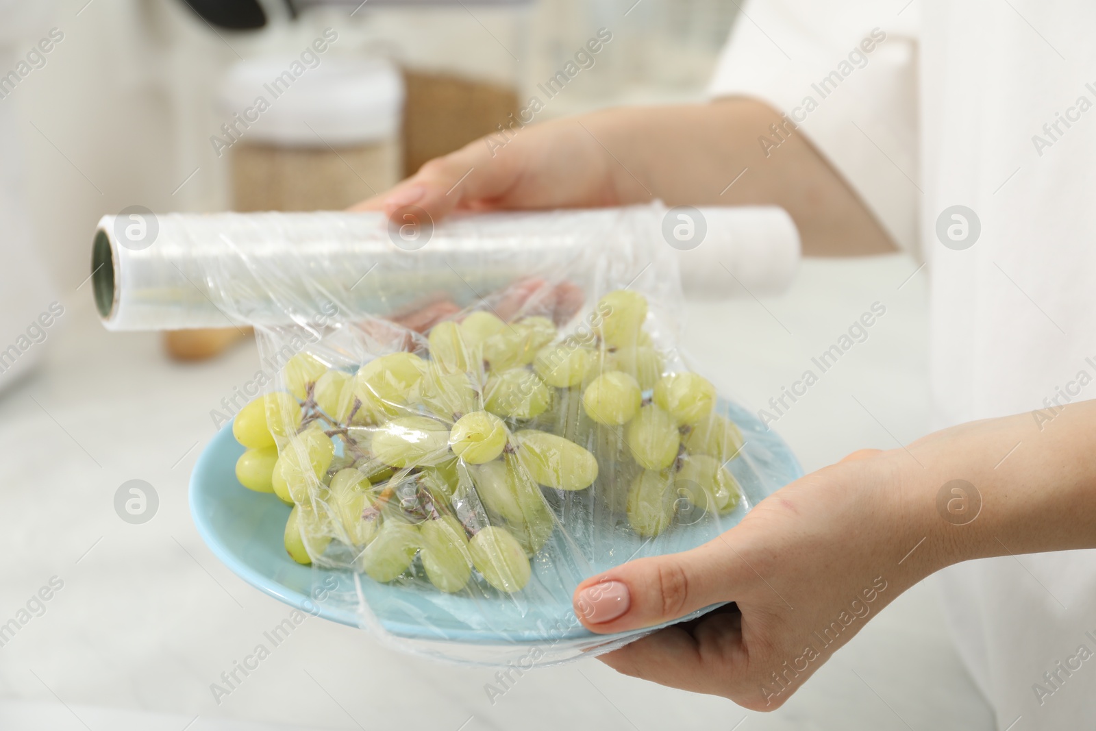 Photo of Woman putting plastic food wrap over plate with grapes in kitchen, closeup