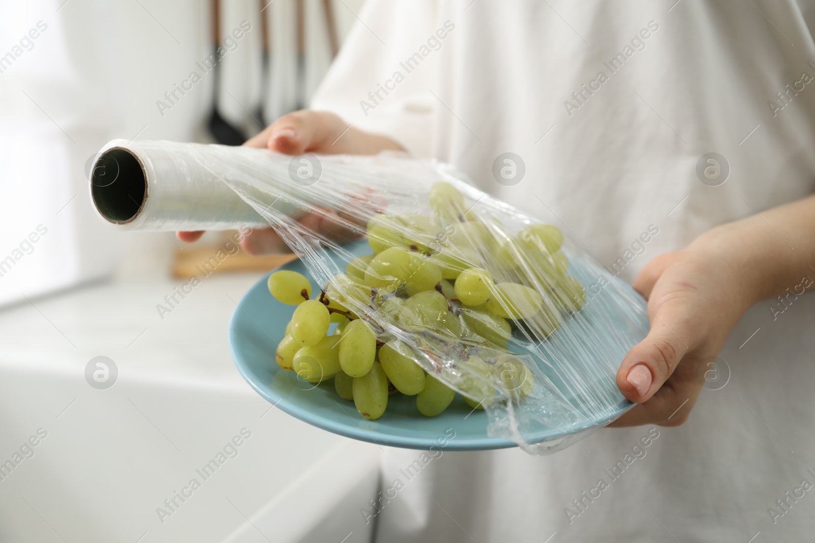 Photo of Woman putting plastic food wrap over plate with grapes in kitchen, closeup