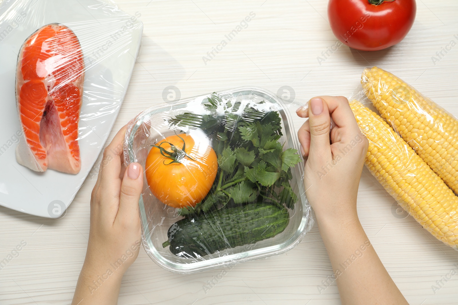 Photo of Woman putting plastic food wrap over glass container with vegetables at white wooden table, top view