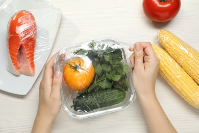 Photo of Woman putting plastic food wrap over glass container with vegetables at white wooden table, top view