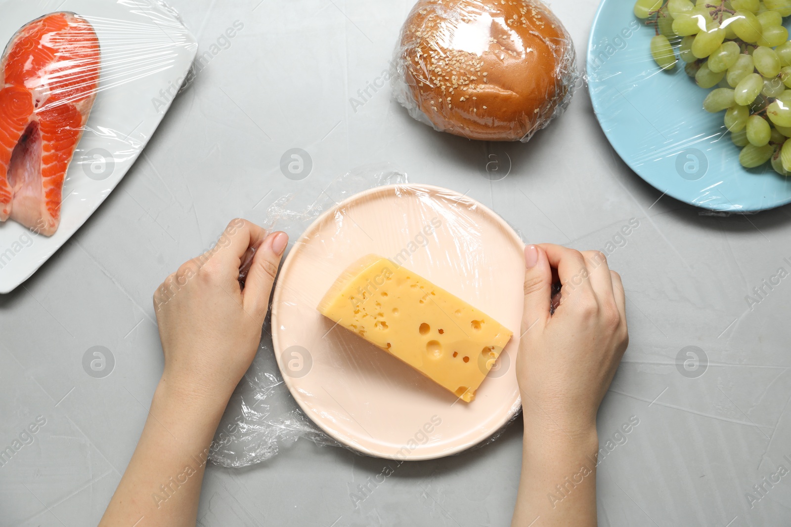 Photo of Woman putting plastic food wrap over plate with cheese at grey table, top view