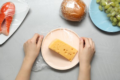 Photo of Woman putting plastic food wrap over plate with cheese at grey table, top view