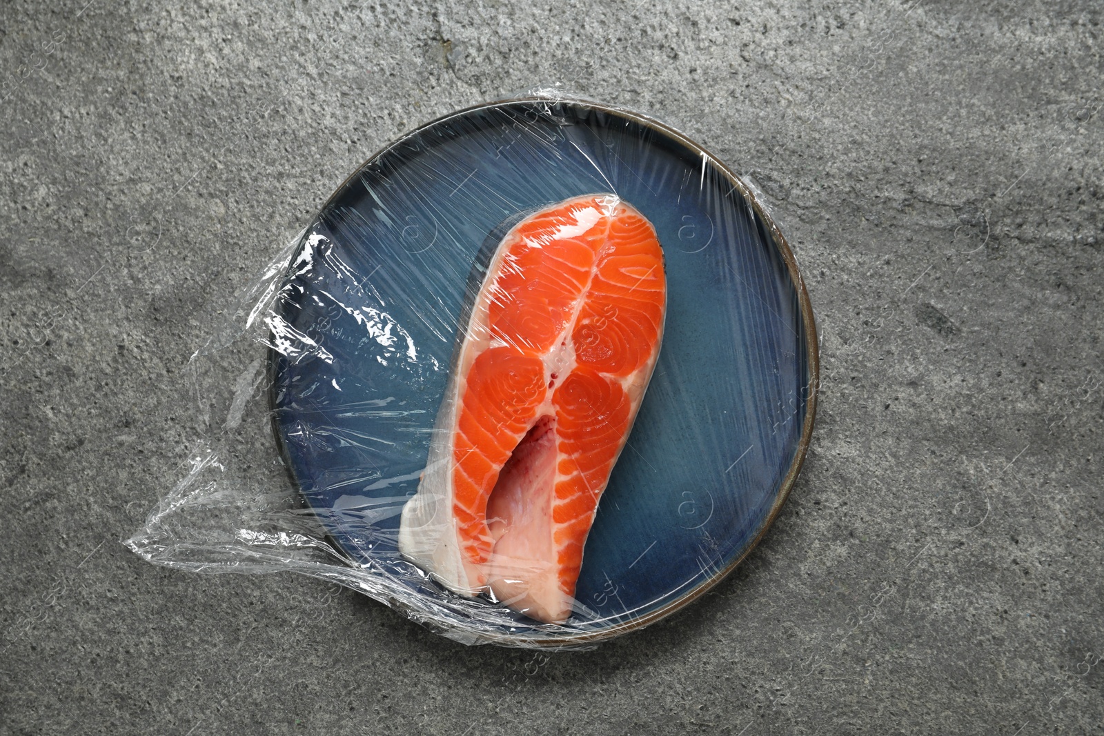 Photo of Plate of salmon with plastic food wrap on grey table, top view