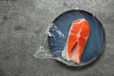 Plate of salmon with plastic food wrap on grey table, top view. Space for text