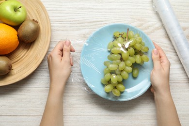 Woman putting plastic food wrap over plate with fresh grapes at wooden table, top view