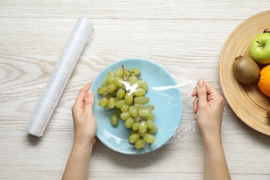 Photo of Woman putting plastic food wrap over plate with fresh grapes at wooden table, top view