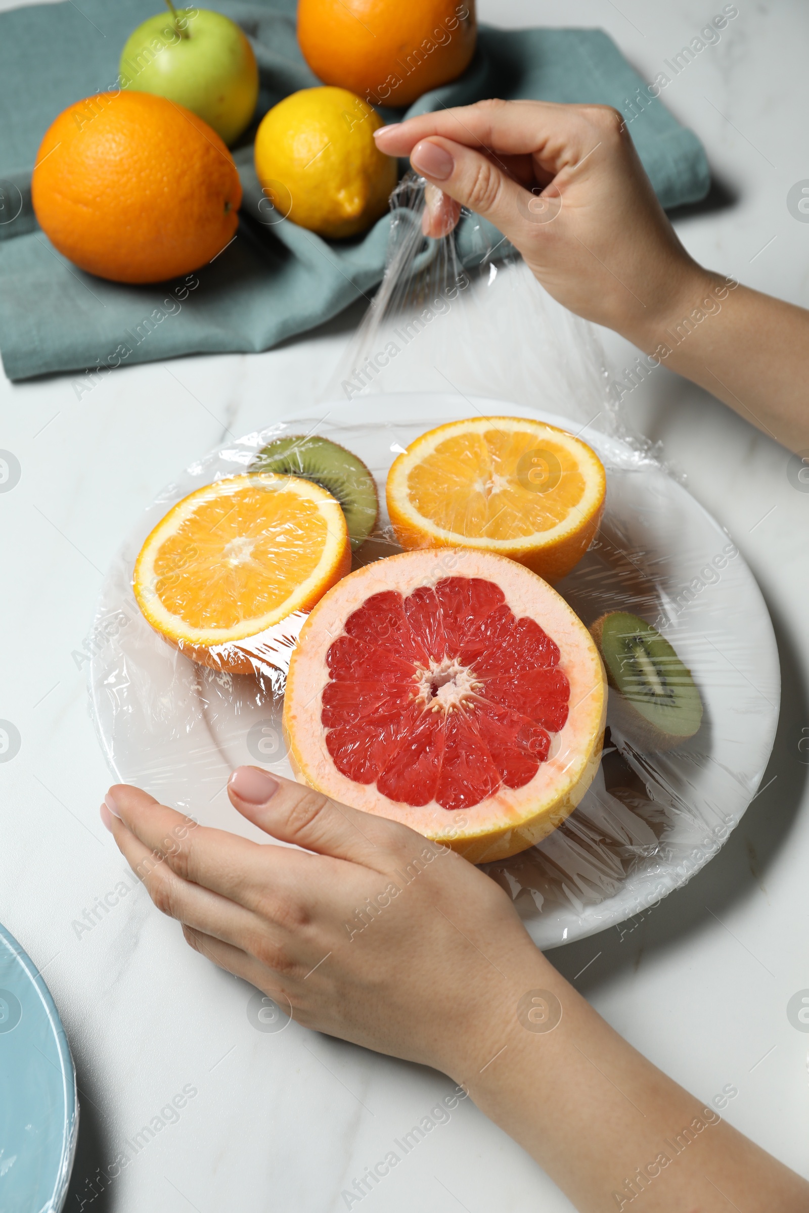 Photo of Woman putting plastic food wrap over plate with fresh fruits at white marble table, closeup