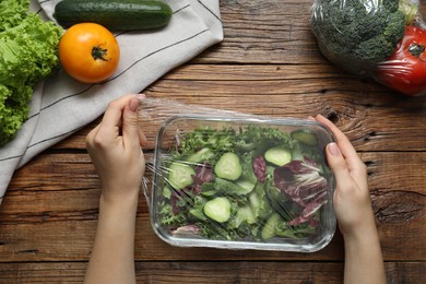 Woman putting plastic food wrap over glass container with salad at wooden table, top view