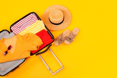 Open suitcase with clothes, wicker hat and shoes on yellow background, flat lay. Space for text