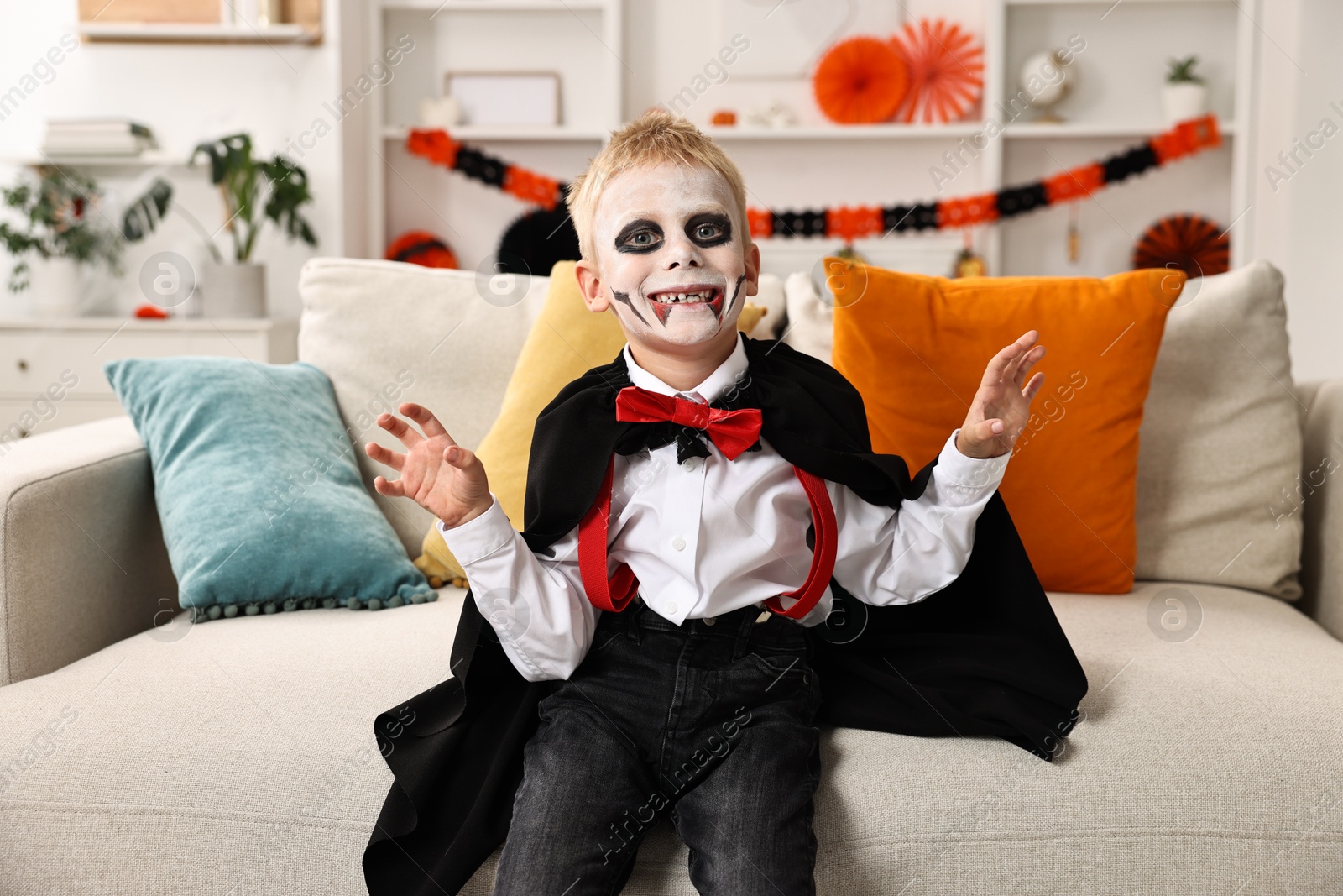 Photo of Funny boy dressed like vampire for Halloween celebration on sofa in room