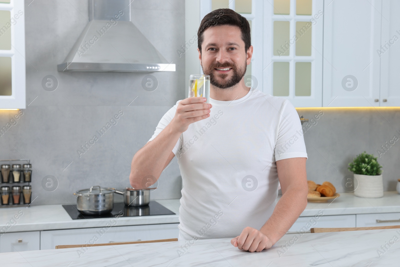 Photo of Happy man holding glass of water with lemon in kitchen