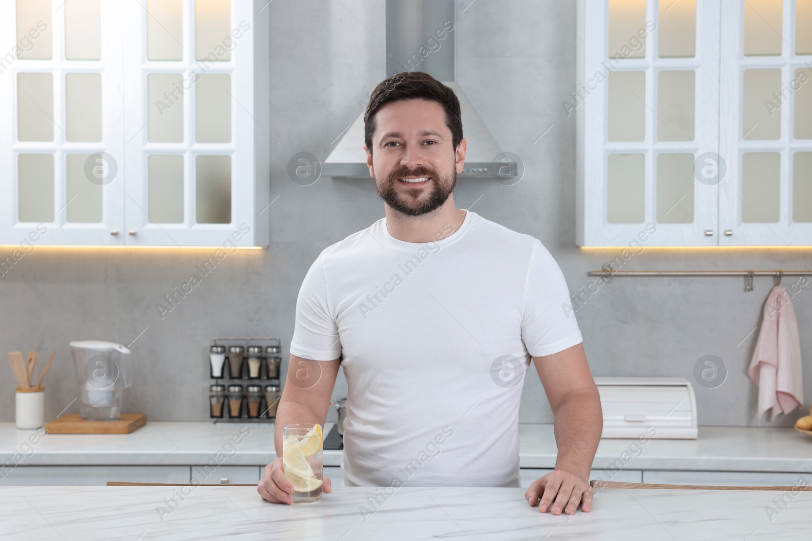 Photo of Happy man holding glass of water with lemon in kitchen