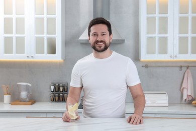 Happy man holding glass of water with lemon in kitchen