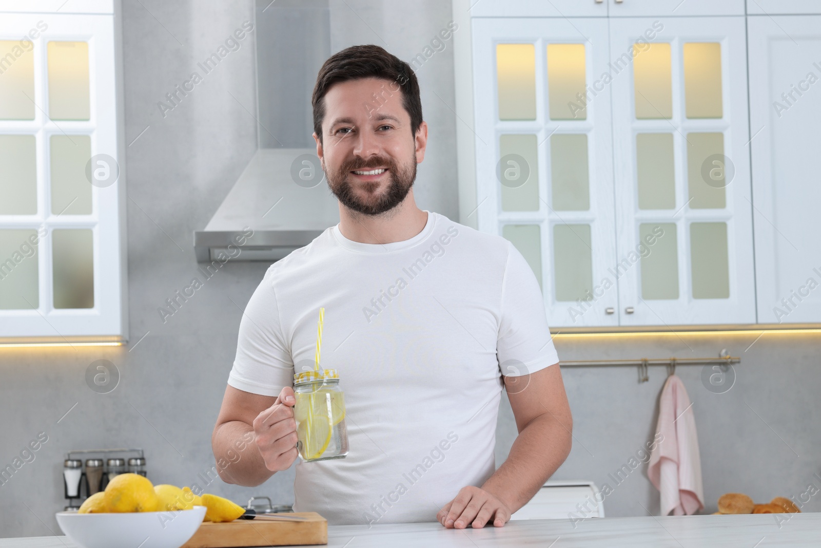 Photo of Happy man holding mason jar of water with lemon in kitchen