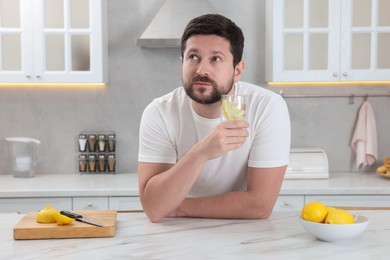 Handsome man holding glass of water with lemon in kitchen