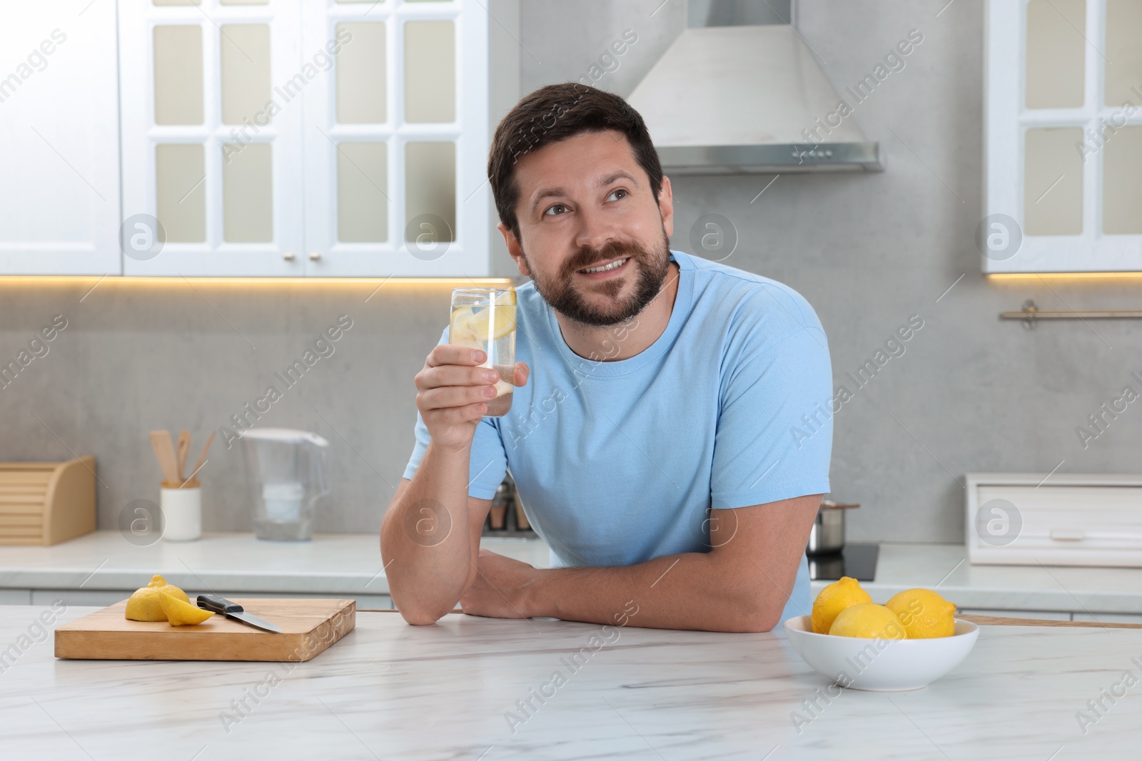 Photo of Happy man holding glass of water with lemon at white marble table in kitchen