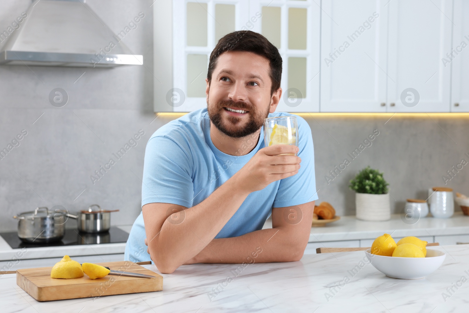 Photo of Happy man holding glass of water with lemon at white marble table in kitchen