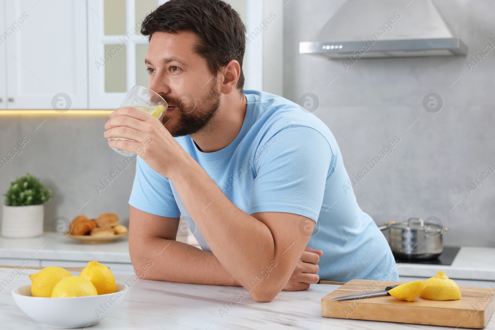 Photo of Handsome man drinking water with lemon at white marble table in kitchen