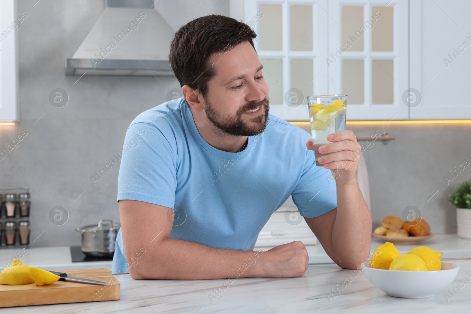 Photo of Happy man holding glass of water with lemon at white marble table in kitchen