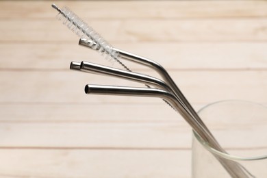 Photo of Metal drinking straws and cleaning brush in glass on wooden table, closeup